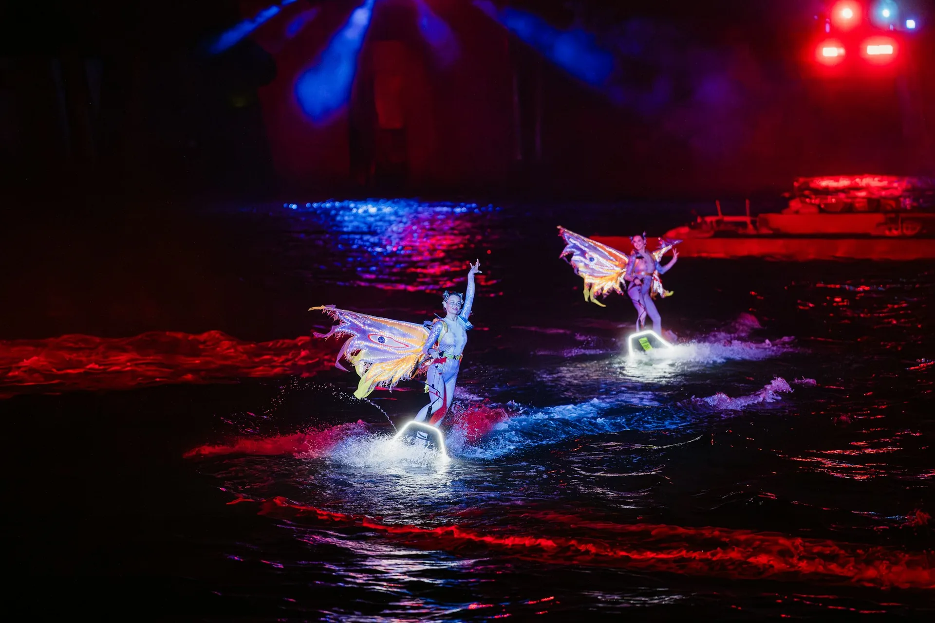 Performers in illuminated costumes skimming across a dark stage pool at night